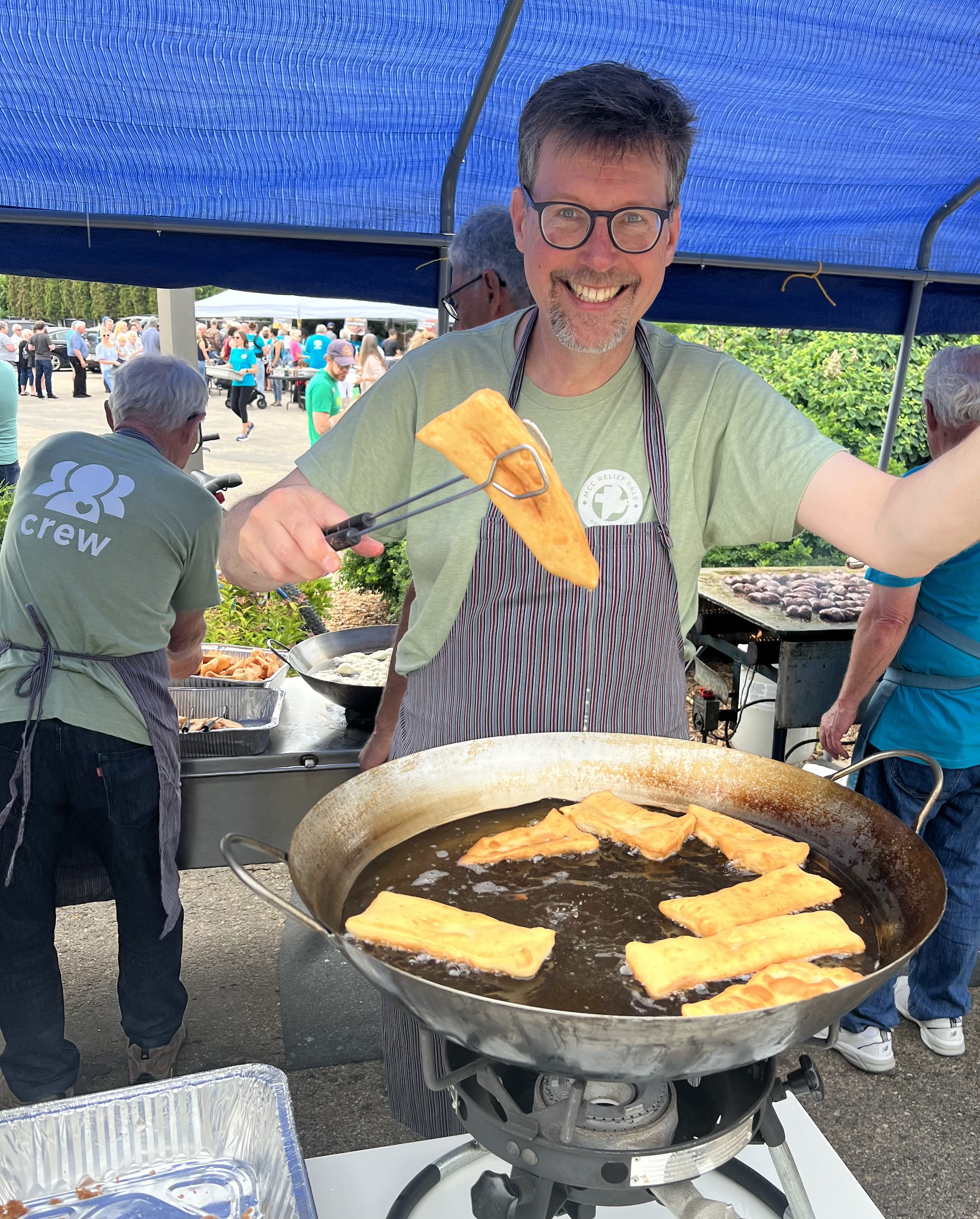A man in an apron fries fish in a large pot of oil under a tent at an outdoor event